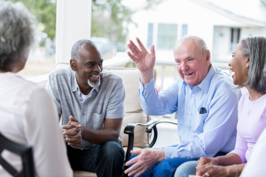A group of seniors sit in a circle together on a front porch and laugh at a joke told by a senior Caucasian man. The man raises his hand.